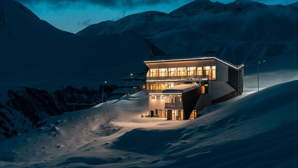 A modern, multi-story building with large windows, situated on a snowy slope with mountains in the background. - Gogi Resort (Kazbegi)