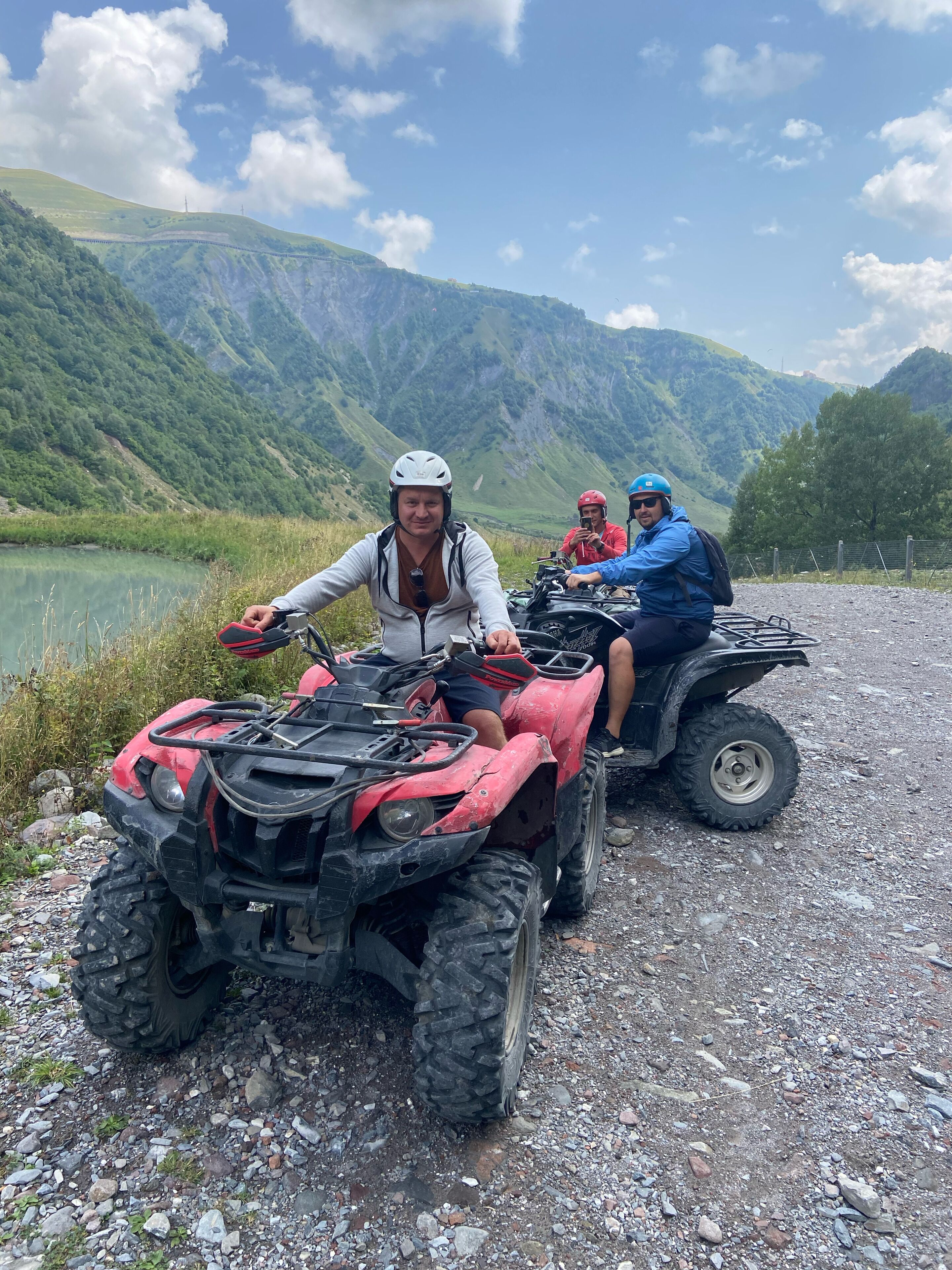 Zwei Personen fahren mit Quads auf einem Schotterweg, im Hintergrund ein See und Berge.