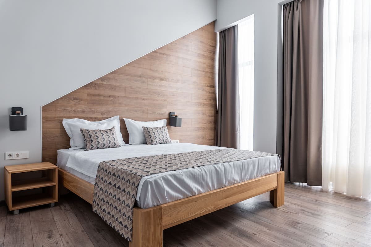 Un dormitorio moderno con cabecera de madera, cama con ropa de cama estampada, mesitas de noche y vistas al exterior a través de amplias ventanas.