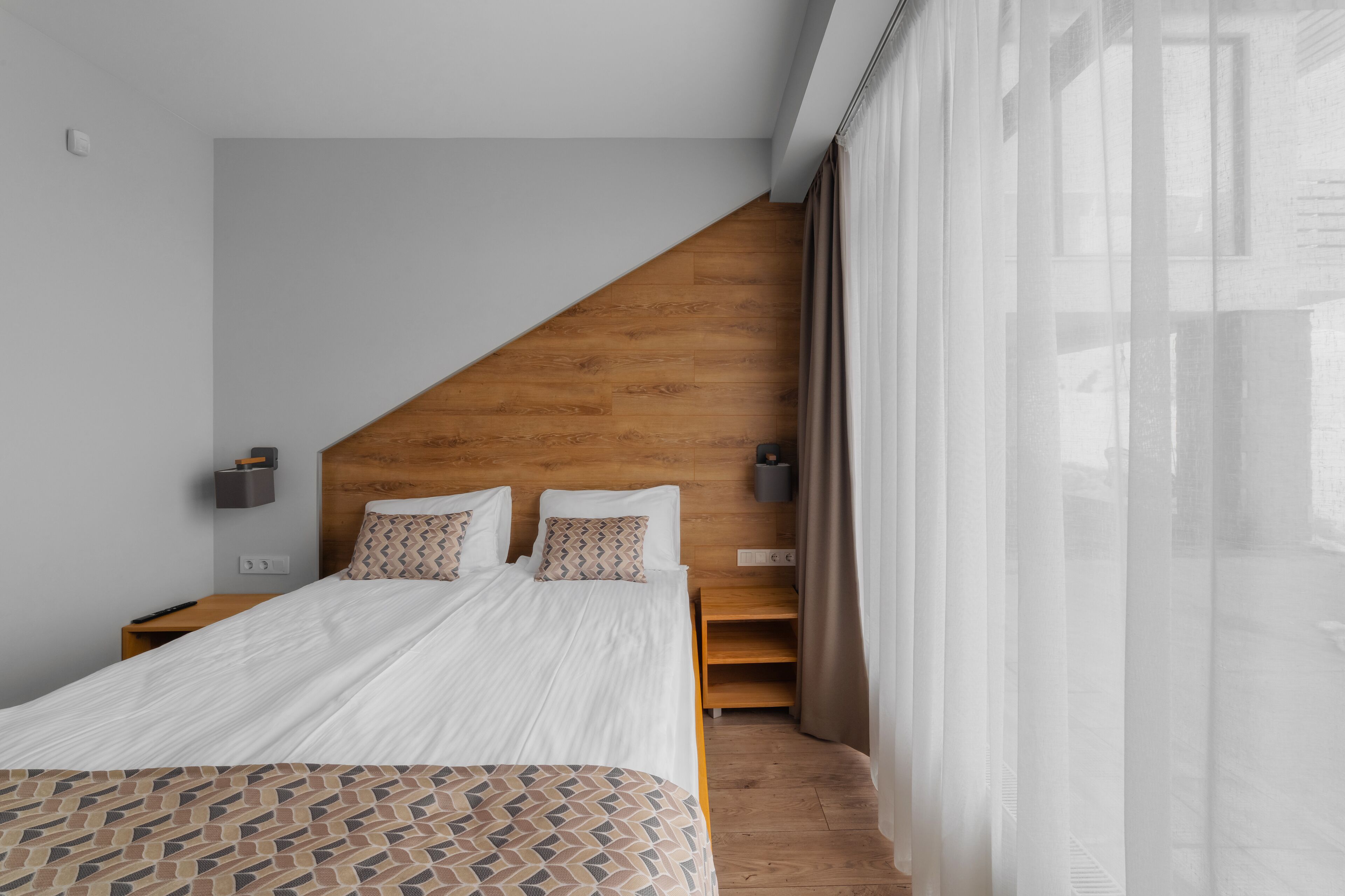 A modern hotel room with a wooden headboard, a large bed with patterned bedding, a nightstand, and a view of the outdoors through sheer curtains.