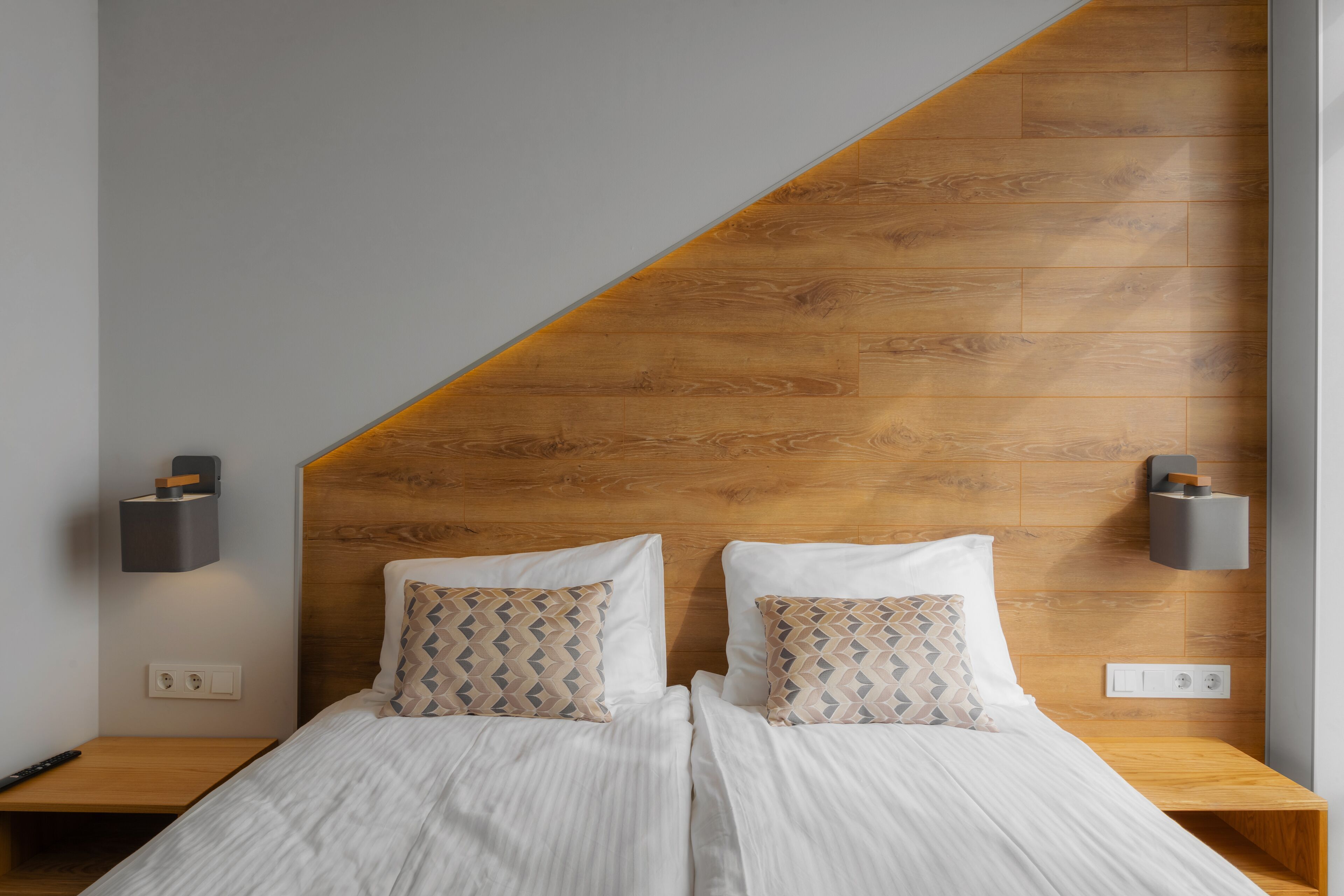 A modern bedroom with a sloped wooden ceiling, two bedside tables with lamps, and patterned pillows.
