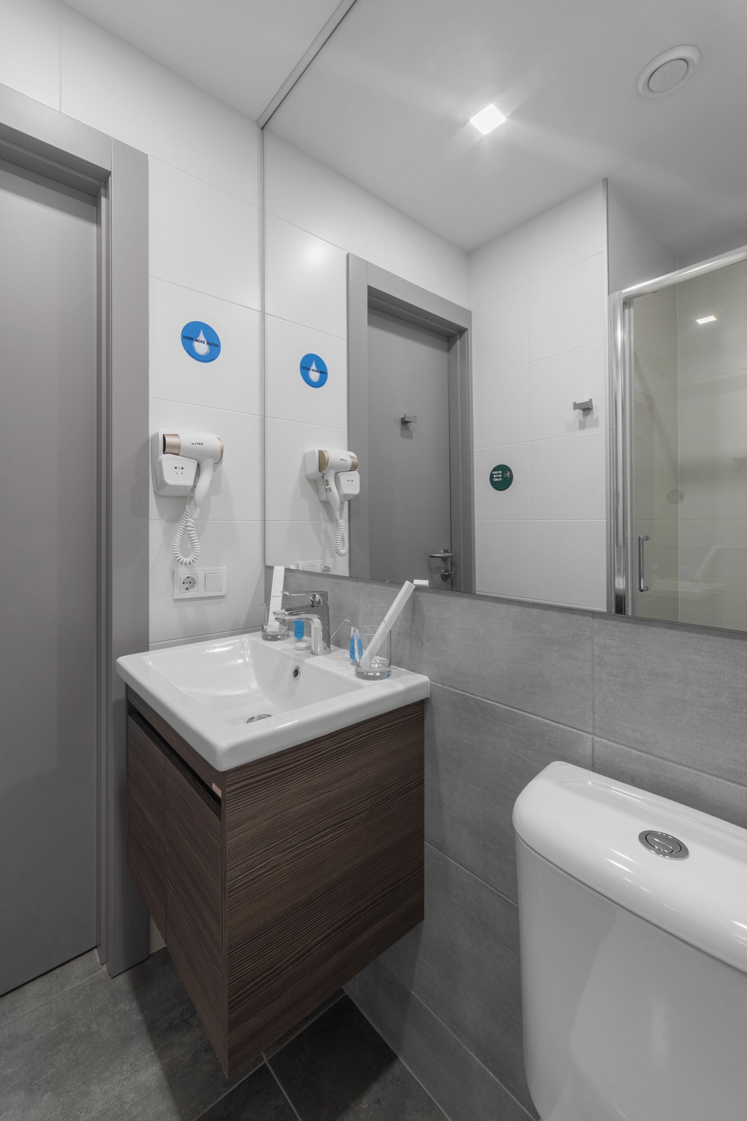 A modern bathroom with a white sink, mirror, and toilet. There are two hair dryers mounted on the wall, and a glass shower enclosure.