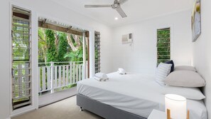 A bedroom with a bed, pillows, a bedside table, a ceiling fan, and a view of greenery through the balcony doors.