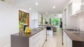 A modern kitchen with a central island, white cabinetry, and a view of a lush garden. - Craven Cottage @ Fullmoon Terraces (PORT DOUGLAS)