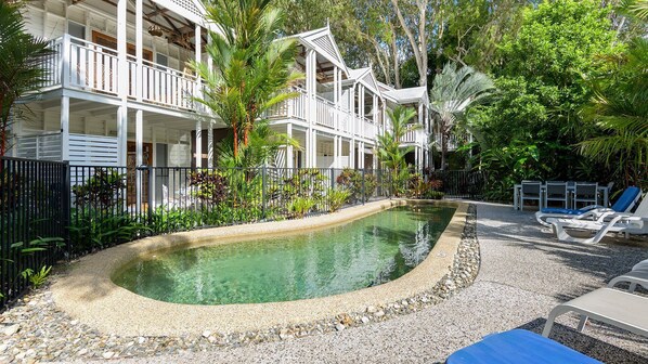 A pool surrounded by a white building with balconies, a gravel path, and lounge chairs.