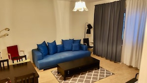 A living room with a blue sofa, a black ottoman, a red chair, a floor lamp, and a window with curtains. - 2 Room Apartment in Hammarby by Stockholm City (Johanneshov)