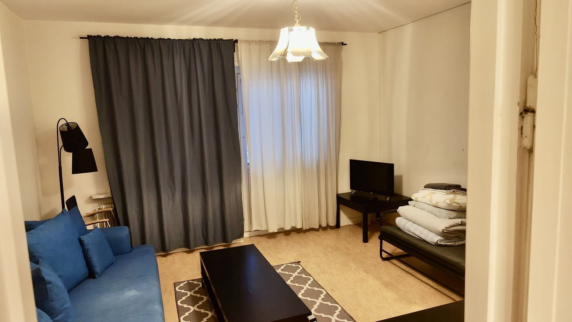Ein Wohnzimmer mit einem blauen Sofa, einem schwarzen Couchtisch, einem Fernseher auf einem Schrank, einer Lampe und einem Fenster mit Vorhängen.