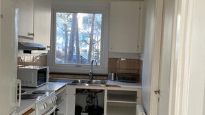 A compact kitchen with white cabinets, a microwave, an oven, a sink, and a window with a view of trees. - 2 Room Apartment in Hammarby by Stockholm City (Johanneshov)