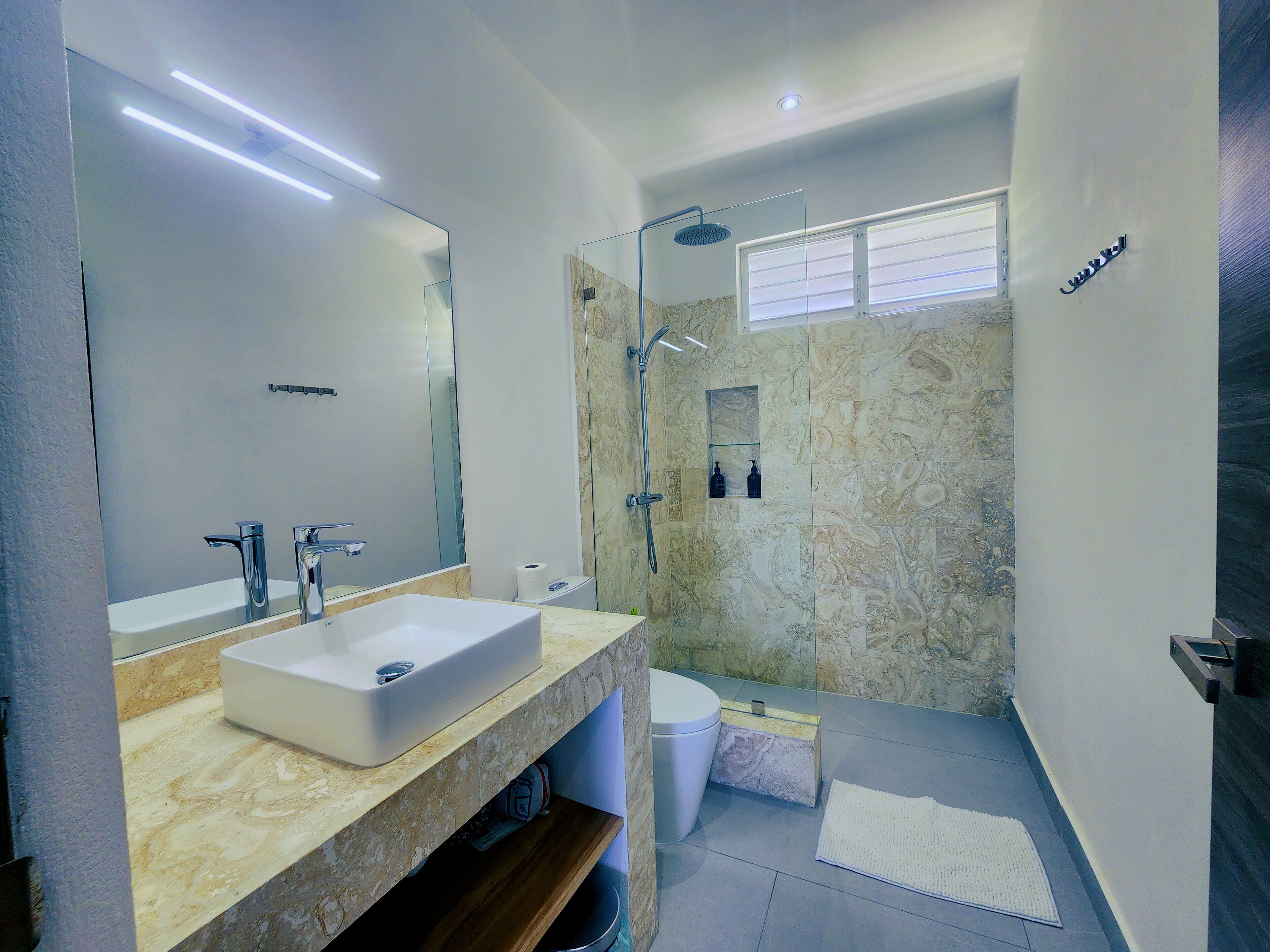 Un baño moderno con un espejo grande, un lavamanos blanco, una ducha con puerta de vidrio y una ventana pequeña.