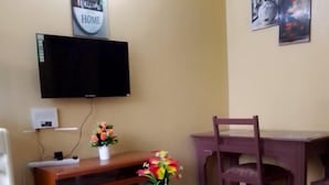 A living room with a TV mounted on the wall, a small table with a vase of flowers, a wooden bench, and a dining table with chairs. - Lux Suites Nairobi House Naivasha (Naivasha)