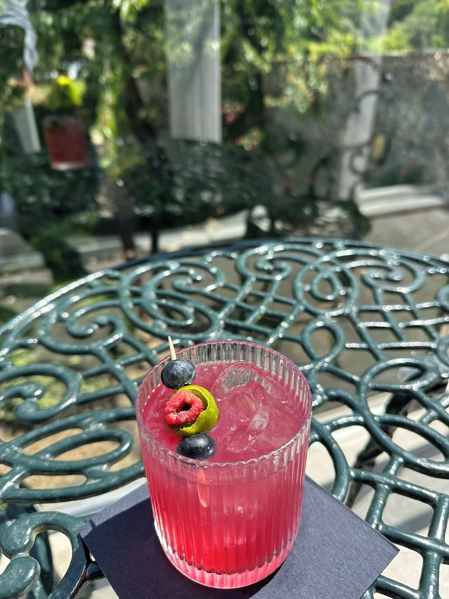Un verre contenant une boisson rouge, décoré d’une framboise et de myrtilles, posé sur une table en métal à l’extérieur.