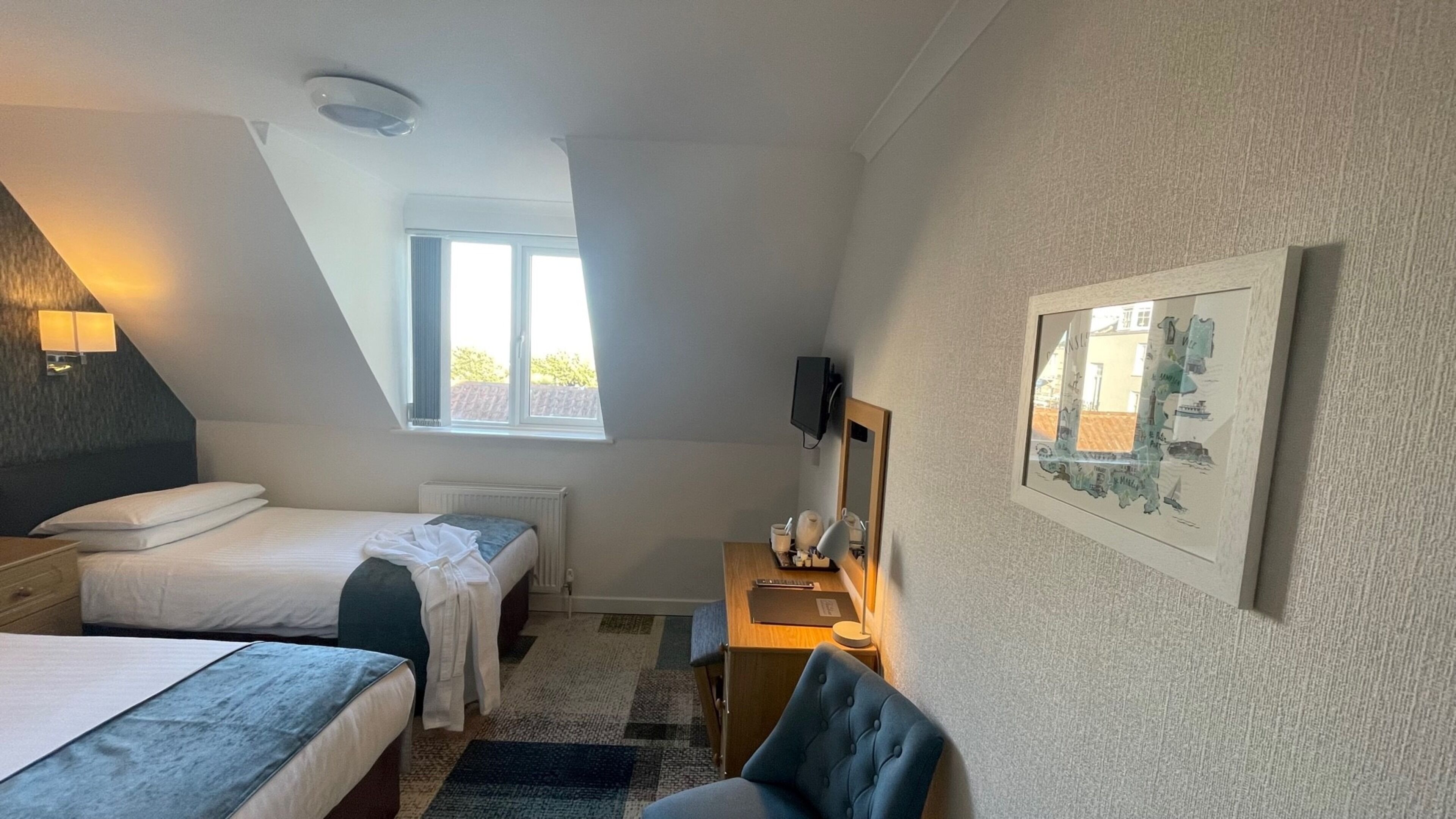 A bedroom with two beds, a desk with a chair, a radiator, a window, a TV, and a framed map on the wall.