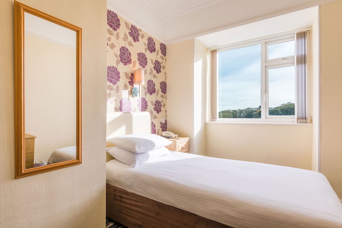 Quarto com cama, mesa de cabeceira, luminária, espelho e janela com vista para o exterior.