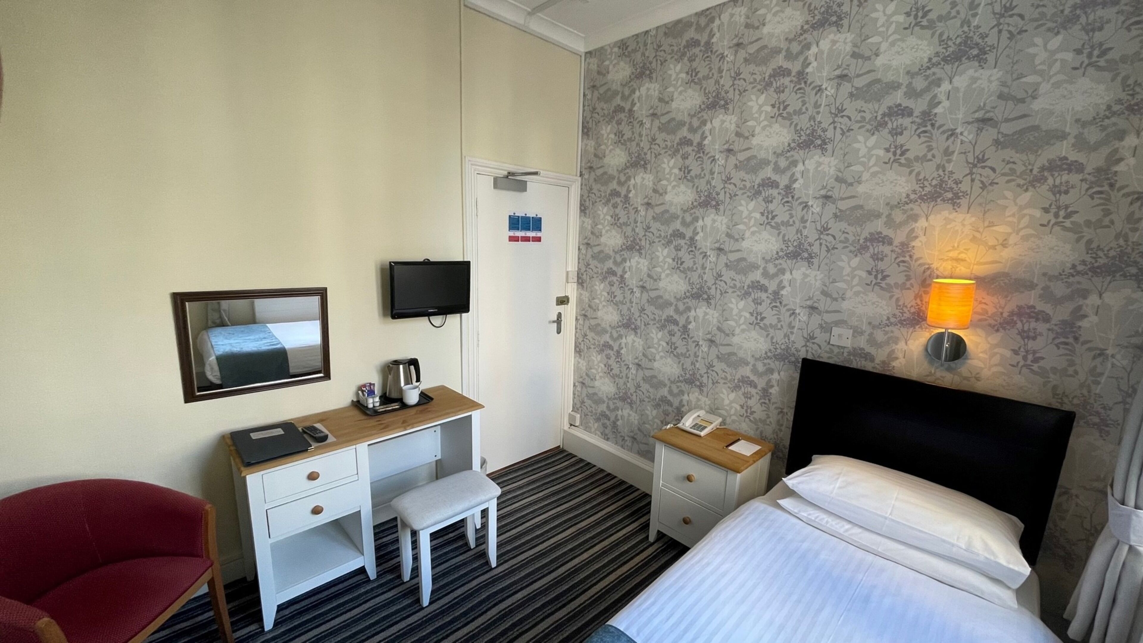 A hotel room with a bed, a desk with a chair, a television, a bedside table with a lamp, and a mirror.