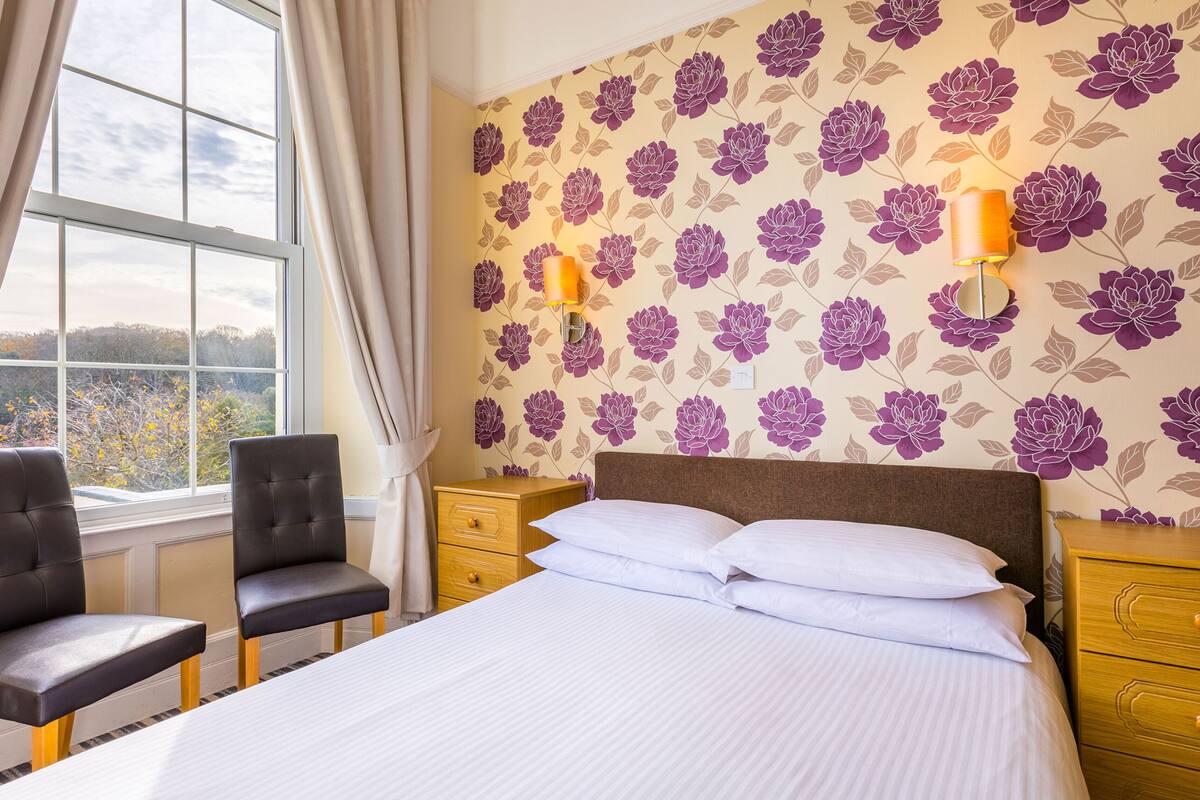 Quarto de hotel com uma cama grande, duas cadeiras, uma mesa de cabeceira e uma janela com cortinas.