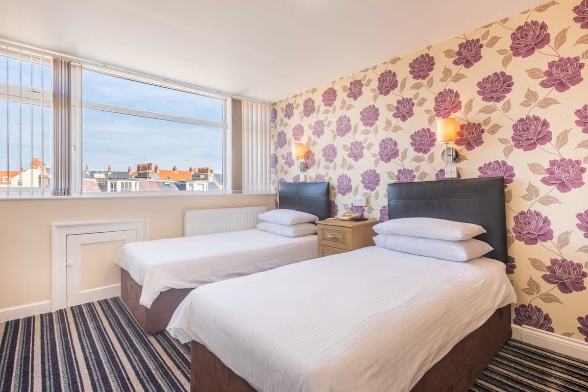 Quarto de hotel com duas camas de solteiro, uma janela grande com vista para edifícios e papel de parede floral.