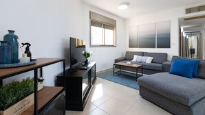 Ein modernes Wohnzimmer mit grauem Sofa, einem Couchtisch, einem Fernseher auf einem Schrank und einem Fenster mit Jalousien.