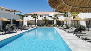 A swimming pool with lounge chairs and umbrellas, surrounded by a building with a tiled roof and palm trees. - Une Vague de Plaisir en Famille (Linguizzetta)