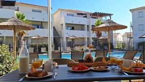 A dining table with breakfast items, a bottle of juice, and a view of a swimming pool and buildings in the background. - Une Vague de Plaisir en Famille (Linguizzetta)