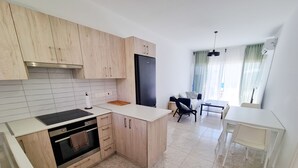 Une cuisine moderne avec des meubles en bois clair, un réfrigérateur noir et un plan de travail blanc. On y trouve un coin repas avec une table et des chaises blanches, ainsi qu’une fenêtre avec des rideaux.