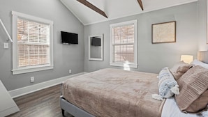 Un dormitorio con cama, dos ventanas con persianas, un televisor montado y una obra de arte enmarcada en la pared.