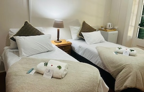 A hotel room with two beds, each with a white duvet and a brown pillow. There is a nightstand with a lamp, a small table with a coffee maker, and a sign indicating the room is for guests.