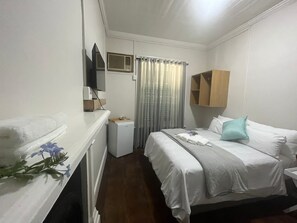 A hotel room with a bed, a small bedside table, a TV, a window with curtains, and a wooden cabinet above the bed.