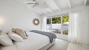 A bedroom with a bed, a ceiling fan, a large window with curtains, and a view of the outdoors. - Sojourn at Sandy Beach - Pool, Beach, Slippery Dip, Style (Sandy Beach)