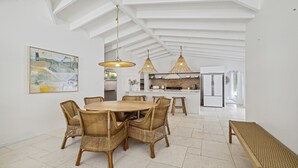A spacious dining area with a round wooden table surrounded by wicker chairs, a kitchen in the background, and a large abstract painting on the wall. - Sojourn at Sandy Beach - Pool, Beach, Slippery Dip, Style (Sandy Beach)