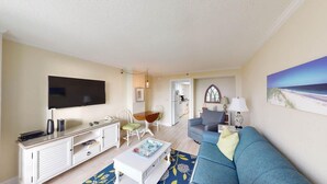 A living room with a TV, white furniture, a blue sofa, and a painting of a beach. - Seaside Bliss Oceanfront1 Bdrm Ste at Bridgewater (Pawleys Island)