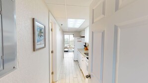 A narrow hallway with white walls and doors, leading to a kitchen area with white cabinets and a window. - Seaside Bliss Oceanfront1 Bdrm Ste at Bridgewater (Pawleys Island)