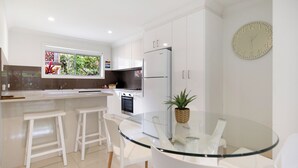 A modern kitchen with white cabinetry, a built-in oven, and a refrigerator. There is a glass dining table with white chairs and a potted plant on the table.