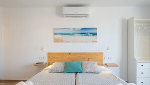 A neatly made double bed with patterned bedding, a wooden headboard, a beach painting above, and a white wardrobe to the right. - Apartamentos Marivent (Felanitx)