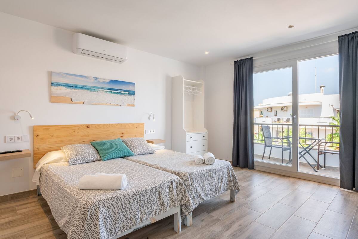 Un dormitorio con cabecera de madera, dos camas con ropa de cama estampada, una mesita de noche, un armario y un balcón con vistas a edificios y vegetación.