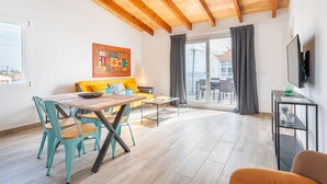 A modern living room with a wooden ceiling, a dining area with turquoise chairs, a sofa with a colorful cushion, and a large window with a view. - Apartamentos Marivent (Felanitx)