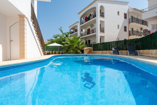 A swimming pool with tiled edges, surrounded by a white building with balconies and a green hedge. - Apartamentos Marivent (Felanitx)