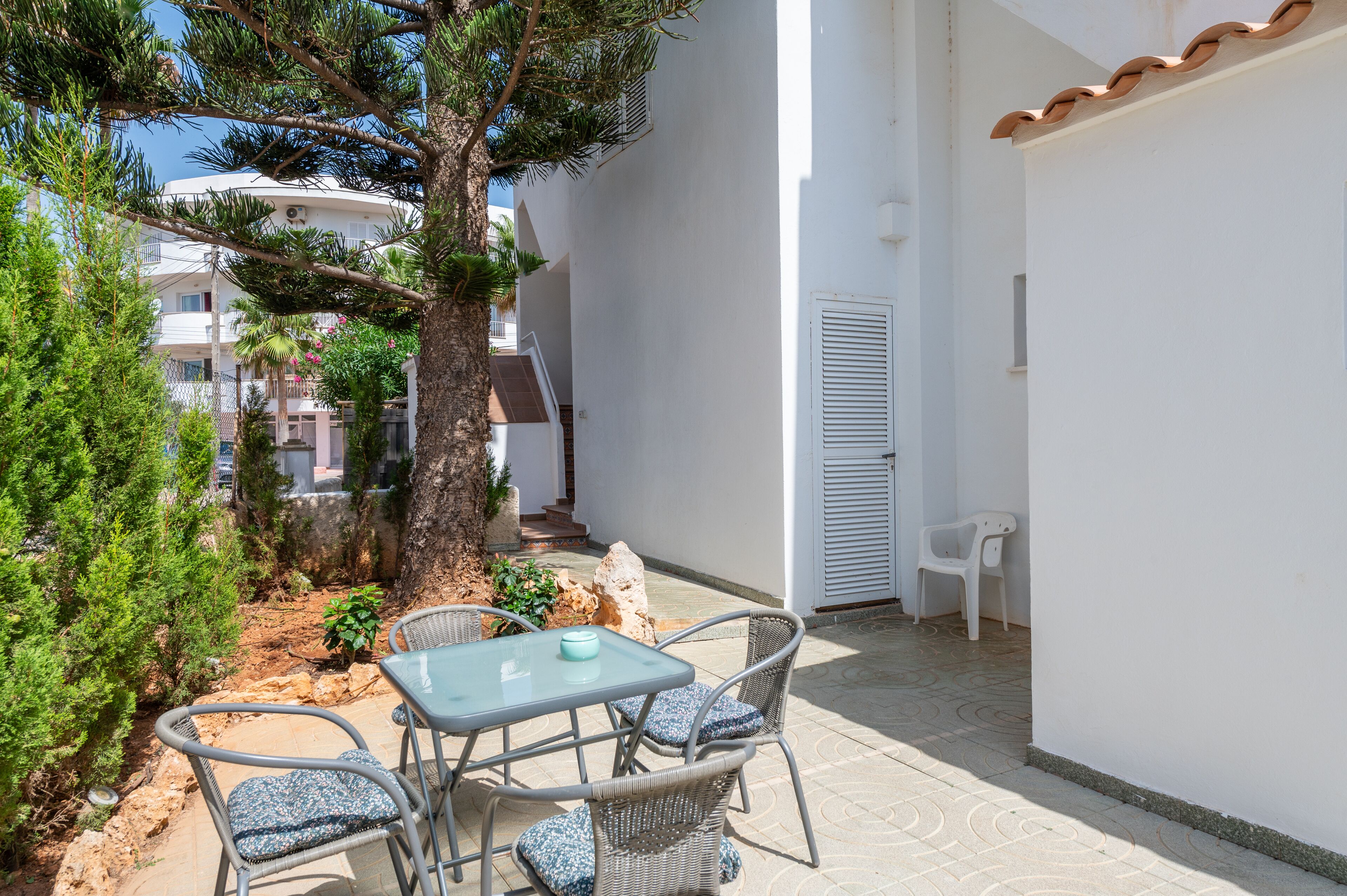 Un patio con una mesa, sillas, un árbol y un edificio blanco con techo de tejas.