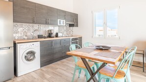 A compact kitchen with grey cabinets, a washing machine, a microwave, and a dining area with a wooden table and teal chairs. - Apartamentos Marivent (Felanitx)