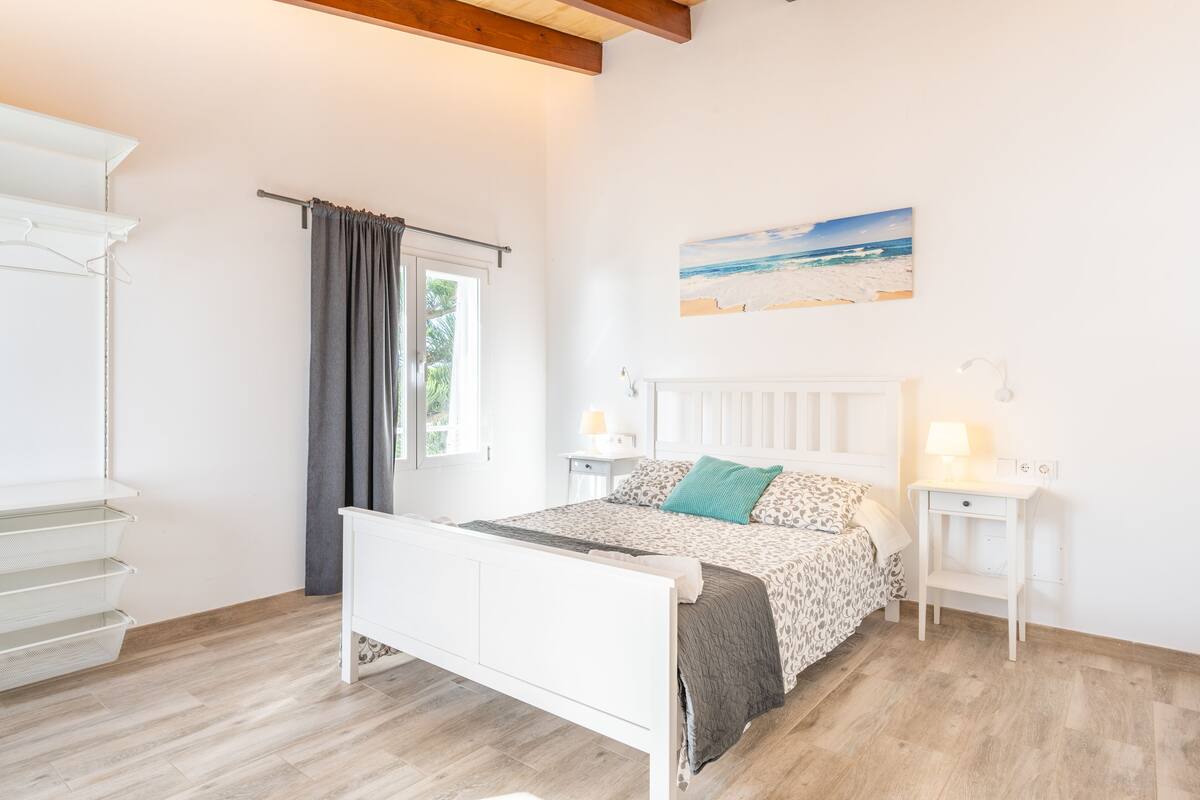 Un dormitorio con cama, mesitas de noche, una ventana con cortinas, un cuadro en la pared y suelo de madera.