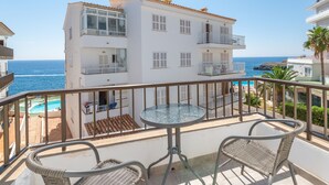 A balcony with a glass table and wicker chairs, overlooking a pool and a building with multiple balconies. - Apartamentos Marivent (Felanitx)