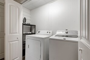 A laundry room with two washing machines and a dryer, a shelf with laundry supplies, and a door. - Marvin`s Gardens: Fenced Yard, Game Room, Pets! (Irondale)