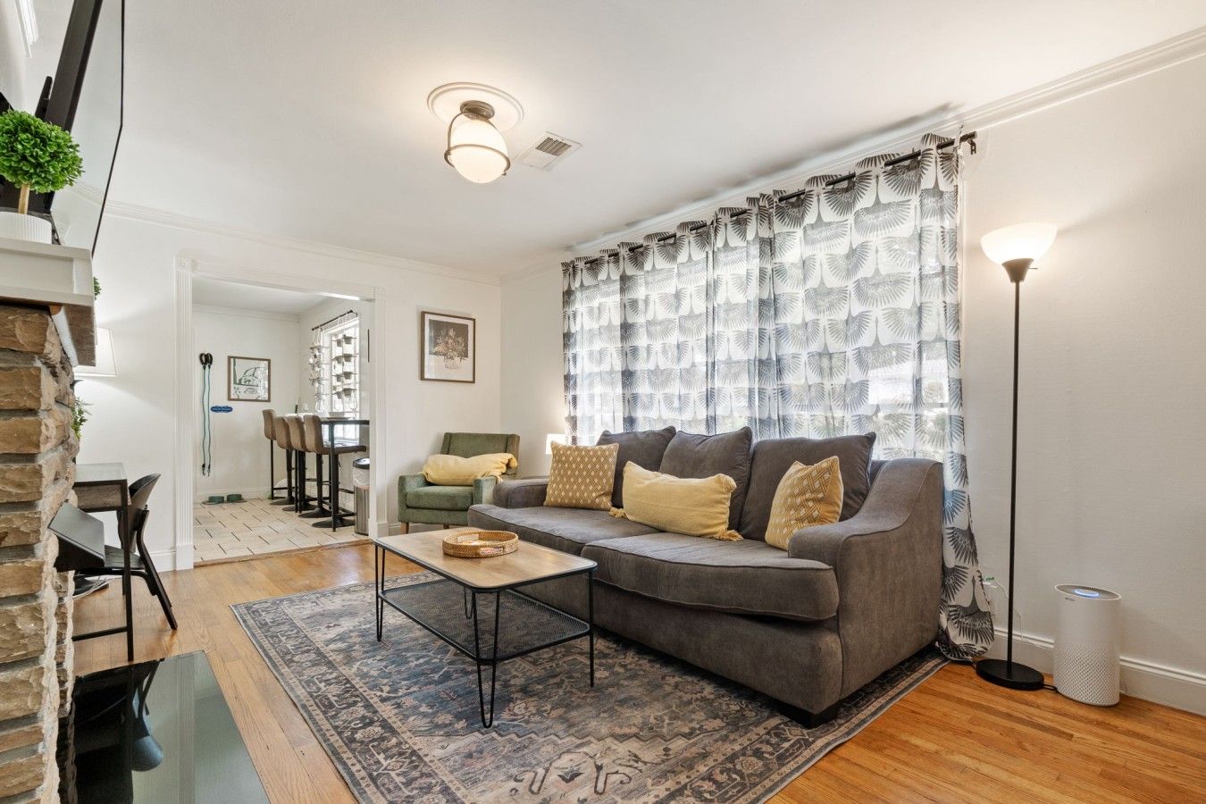 Ein Wohnzimmer mit grauem Sofa, gemusterten Vorhängen, einer Stehlampe und einem Essbereich im Hintergrund.