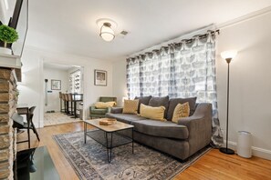 Una sala de estar con un sofá gris, cortinas estampadas, una lámpara de pie y un comedor al fondo.
