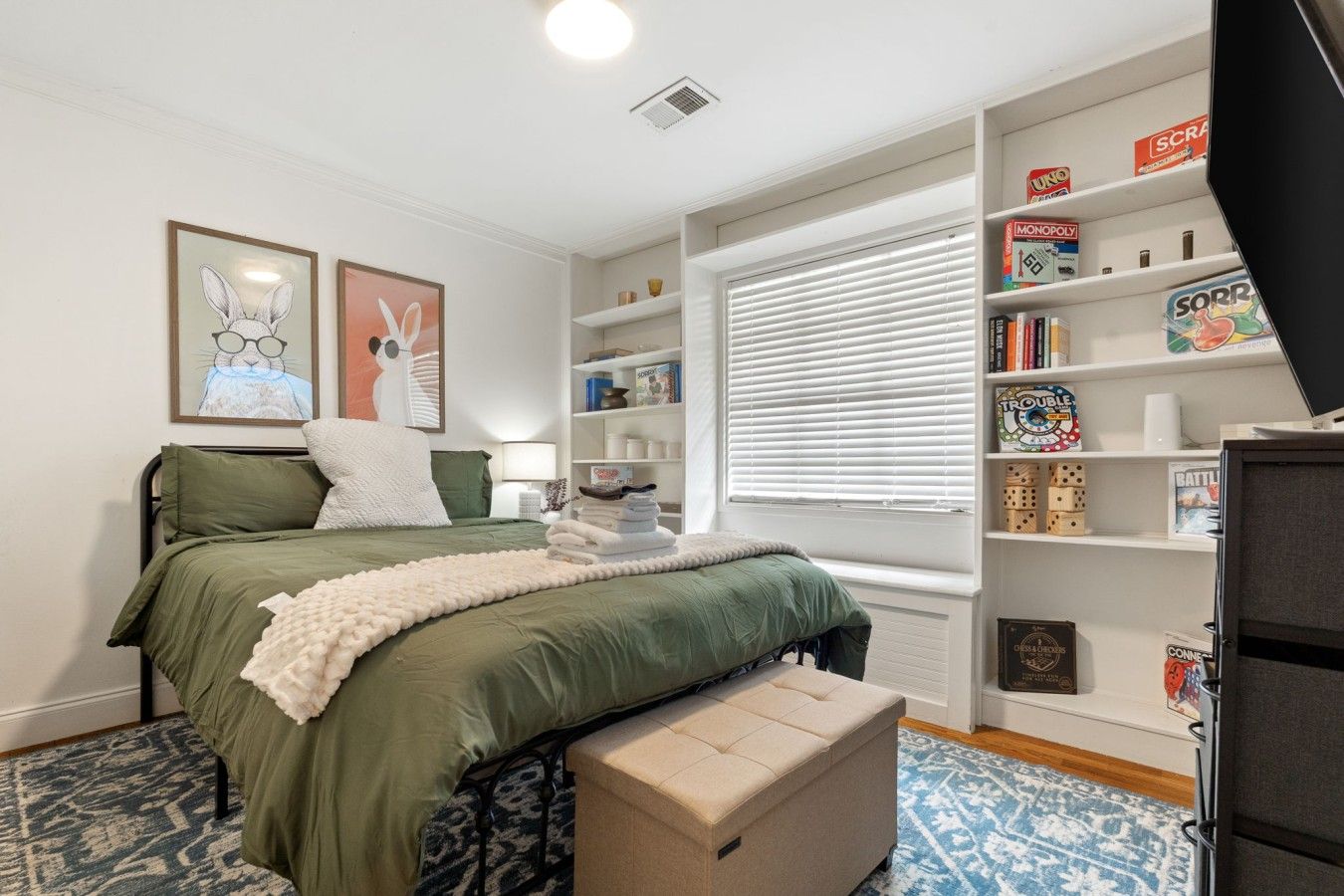Un dormitorio con cama, una ventana con persianas, una estantería con juegos de mesa y una mesita con lámpara.