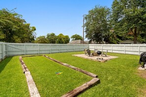Un patio trasero con césped, una terraza de madera y una zona de barbacoa con sillas y una parrilla.
