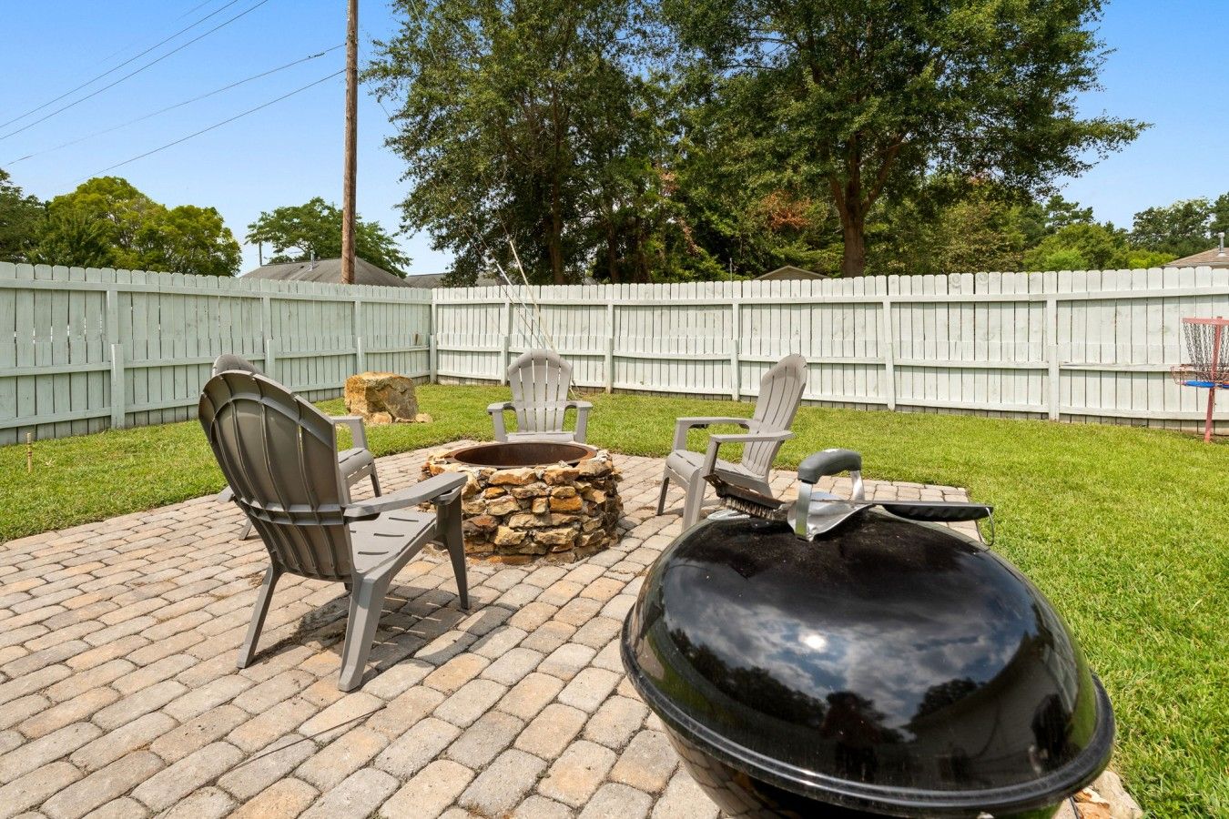 Un patio trasero con un hogar de piedra, dos sillas Adirondack y una parrilla negra grande.