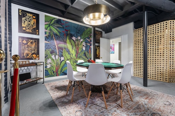 A room with a green felt table, white chairs, a red bar cart, and a tropical mural on the wall.