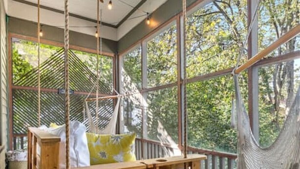 A hammock chair suspended from the ceiling, a wooden bed with pillows, and a view of trees through large windows. - The Glen Iris Getaway (Birmingham)