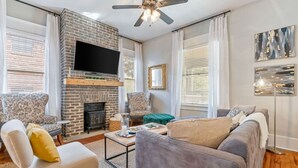 A living room with a brick fireplace, a flat-screen TV, two armchairs, a sofa, and a ceiling fan. - The Glen Iris Getaway (Birmingham)
