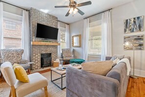 A living room with a brick fireplace, a flat-screen TV, two armchairs, a sofa, and a ceiling fan.